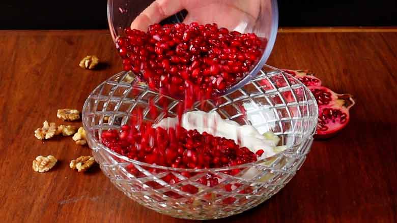 A hand is seen tipping pomegranate arils from a glass bowl over yogurt and cucumbers in large cut-glass bowl. California walnuts kernels and an open pomegranate lie on the wooden counter in the background - Konkani Cucumber and Pomegranate Salad Recipe